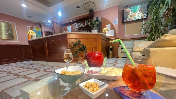 Un área de bar con suelo a cuadros, una mesa de vidrio con aperitivos, una manzana roja, una copa de vino y un televisor montado en la pared.