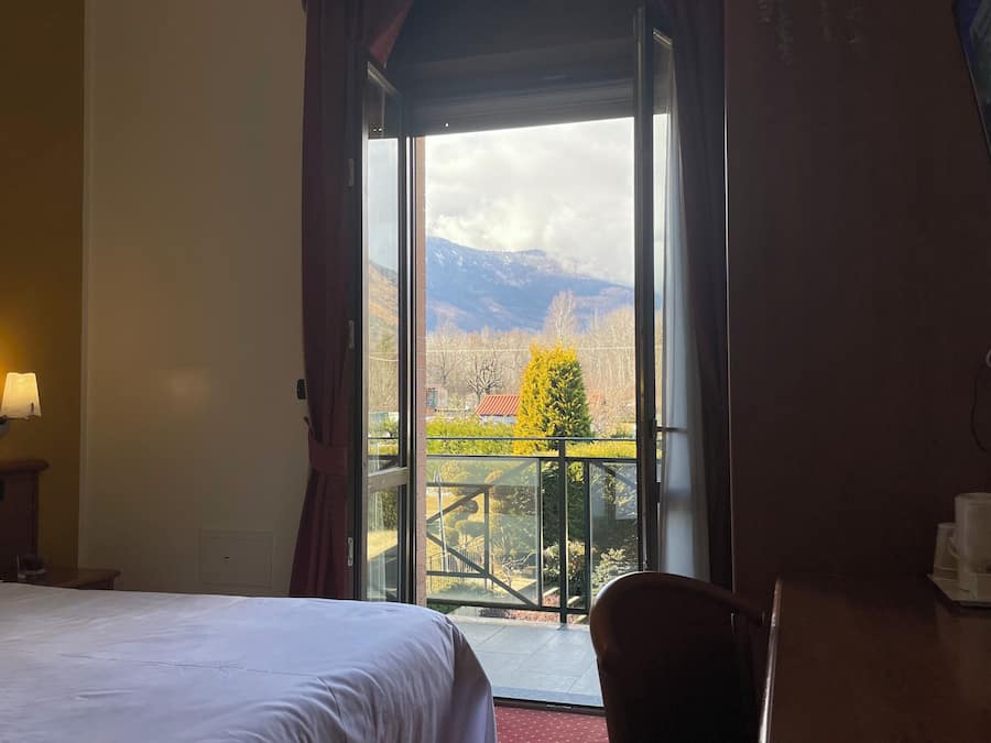 Quarto de hotel com cama, escrivaninha, cadeira, janela com vista para montanhas e varanda.