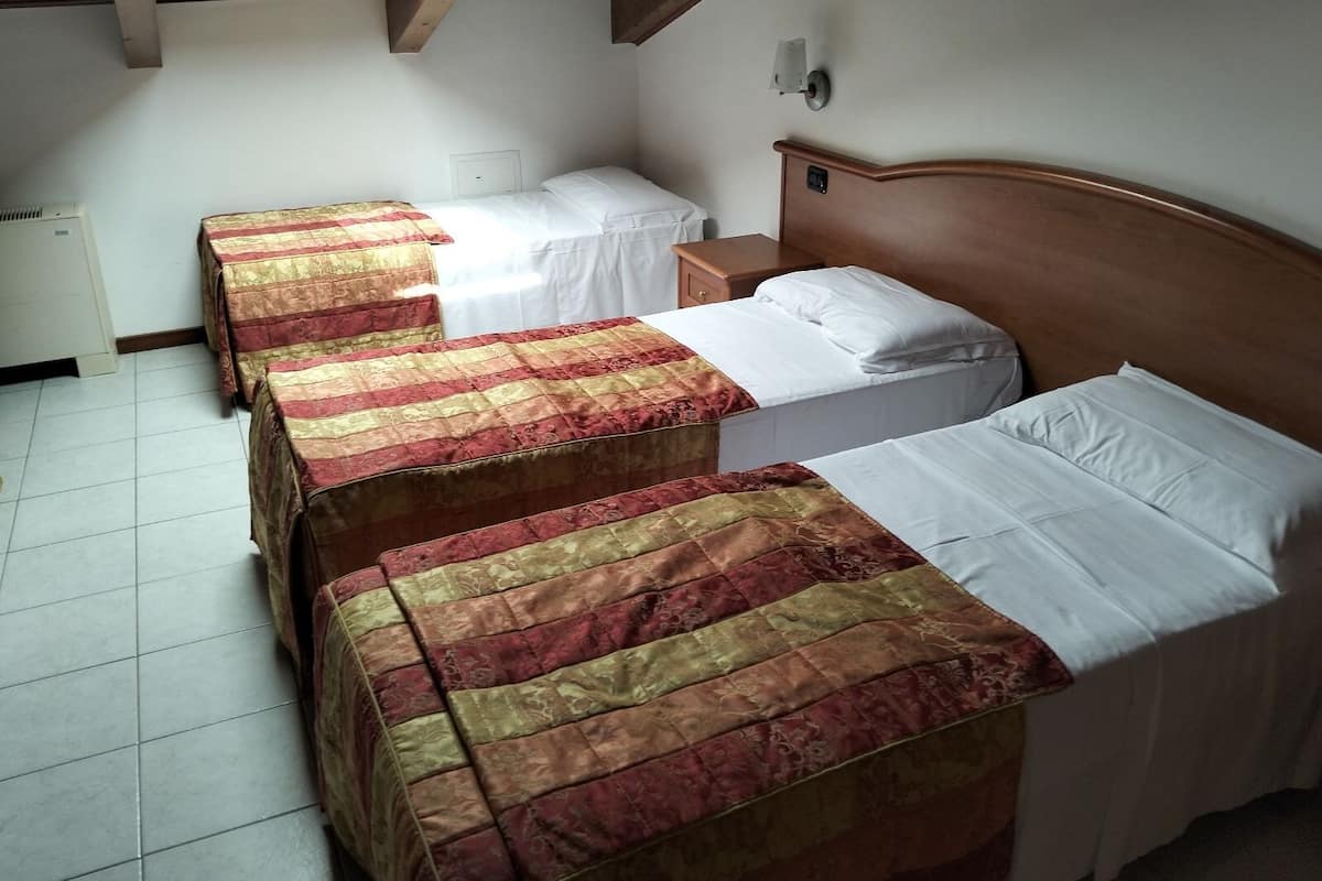 Ein Zimmer mit drei Betten, jedes mit unterschiedlicher Bettwäsche. Das Bett rechts hat ein hölzernes Kopfteil, eine weiße Wand und ein Fenster mit Tageslicht.