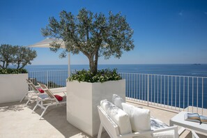 A balcony with white lounge chairs, a white umbrella, and a tree planter with flowers. - Casa Angelina (Praiano)