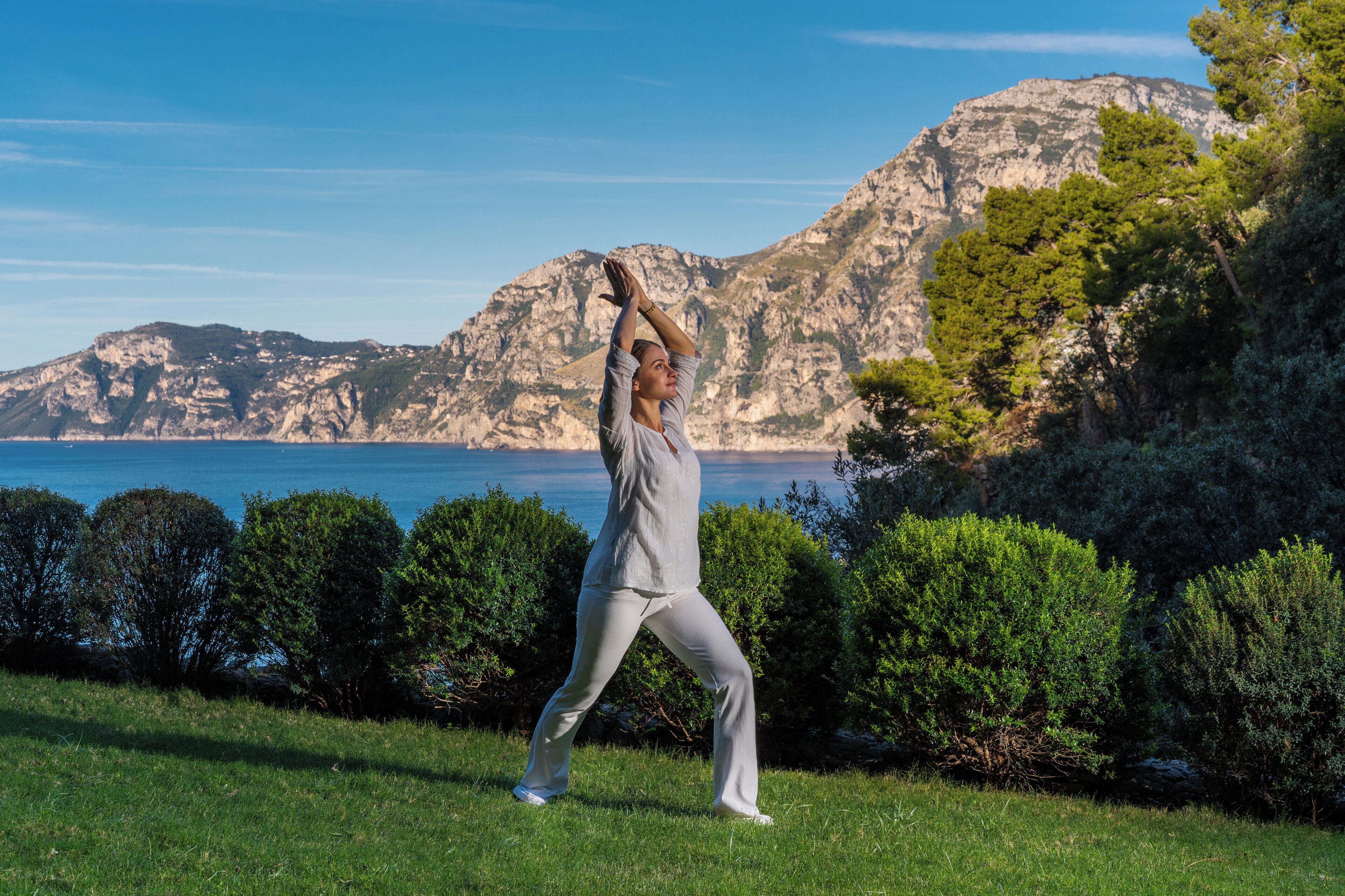 Una persona realizando una postura de yoga en una zona cubierta de hierba, con montañas y un cuerpo de agua al fondo.