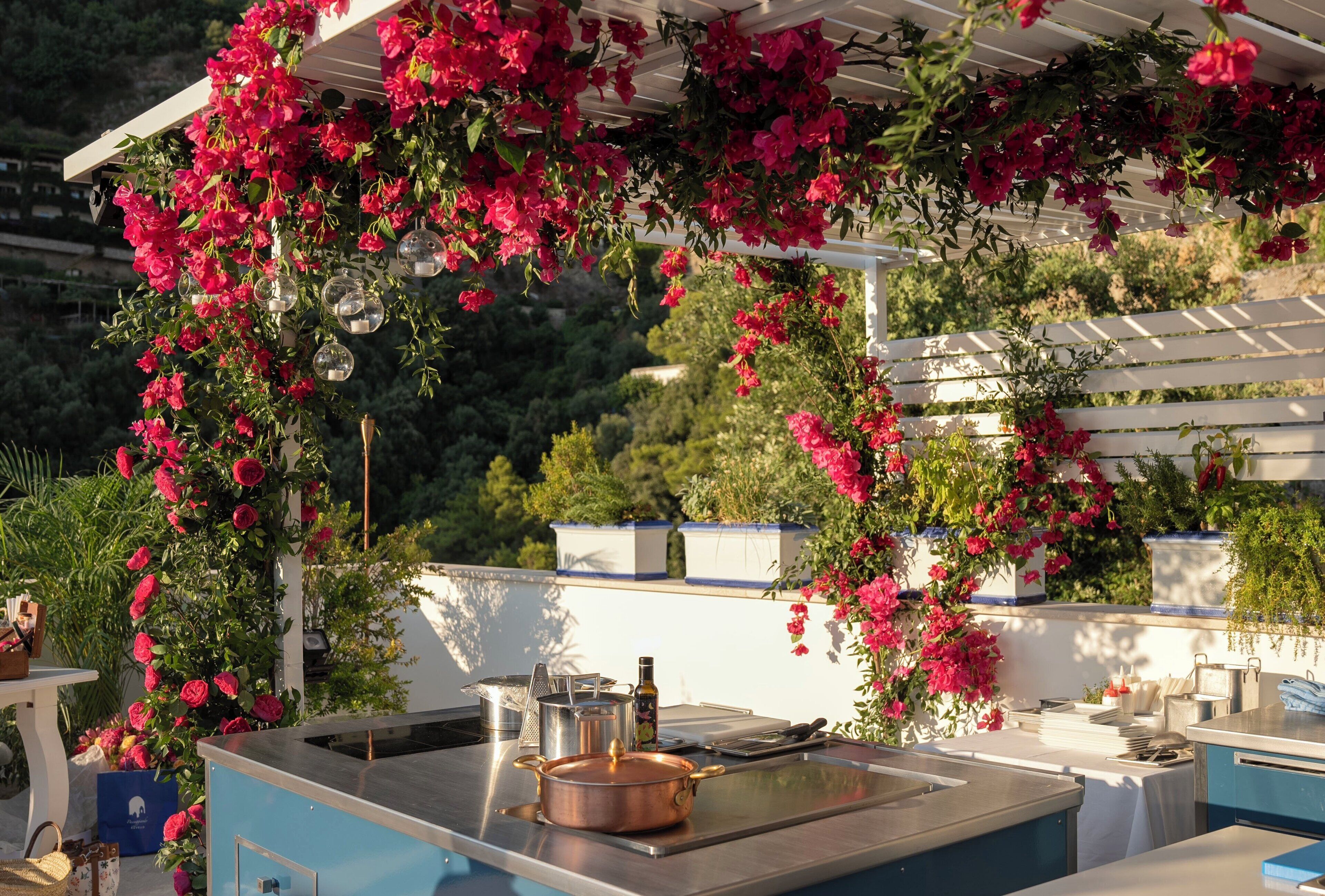 Zona de cocina exterior con encimera azul, una olla de cobre y flores rojas colgantes.
