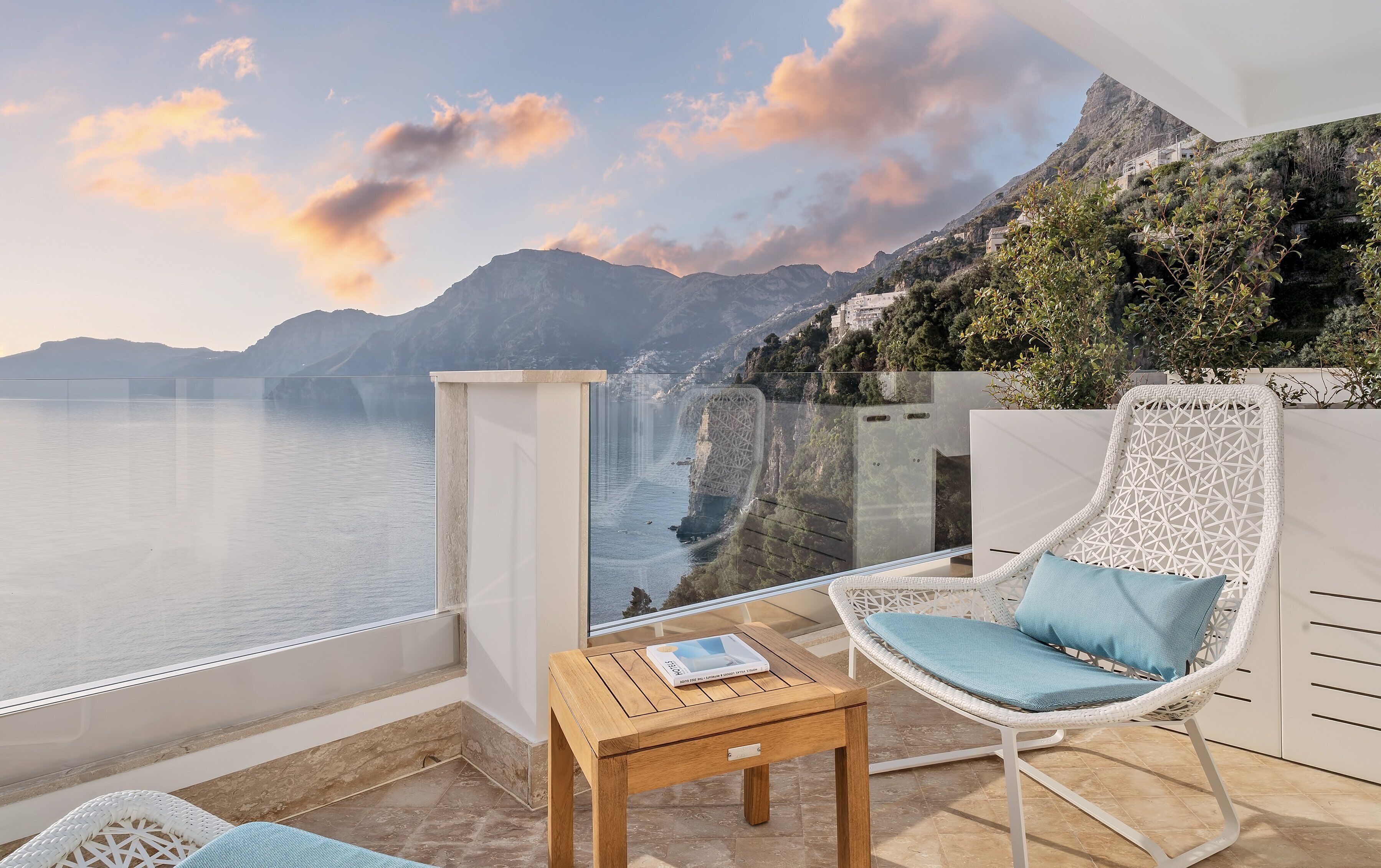 A balcony with a view of the sea and mountains, a wicker chair with a cushion, a small wooden table, and a glass partition.