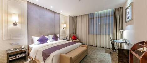 Quarto de hotel com uma cama grande, uma escrivaninha, uma cadeira, uma mesinha de cabeceira e uma janela com cortinas.