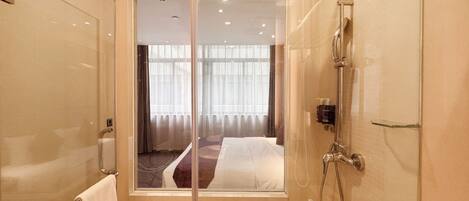 A modern bathroom with a glass shower enclosure, a visible bed with curtains, and a towel rack.