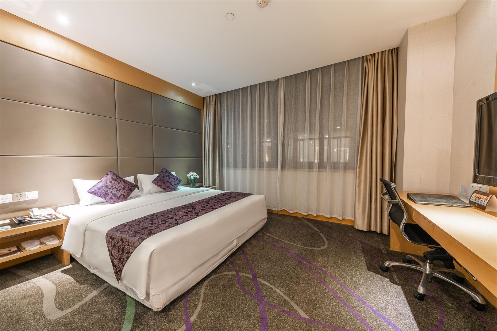 A hotel room with a large bed, a desk with a chair, a TV, and a window with curtains.