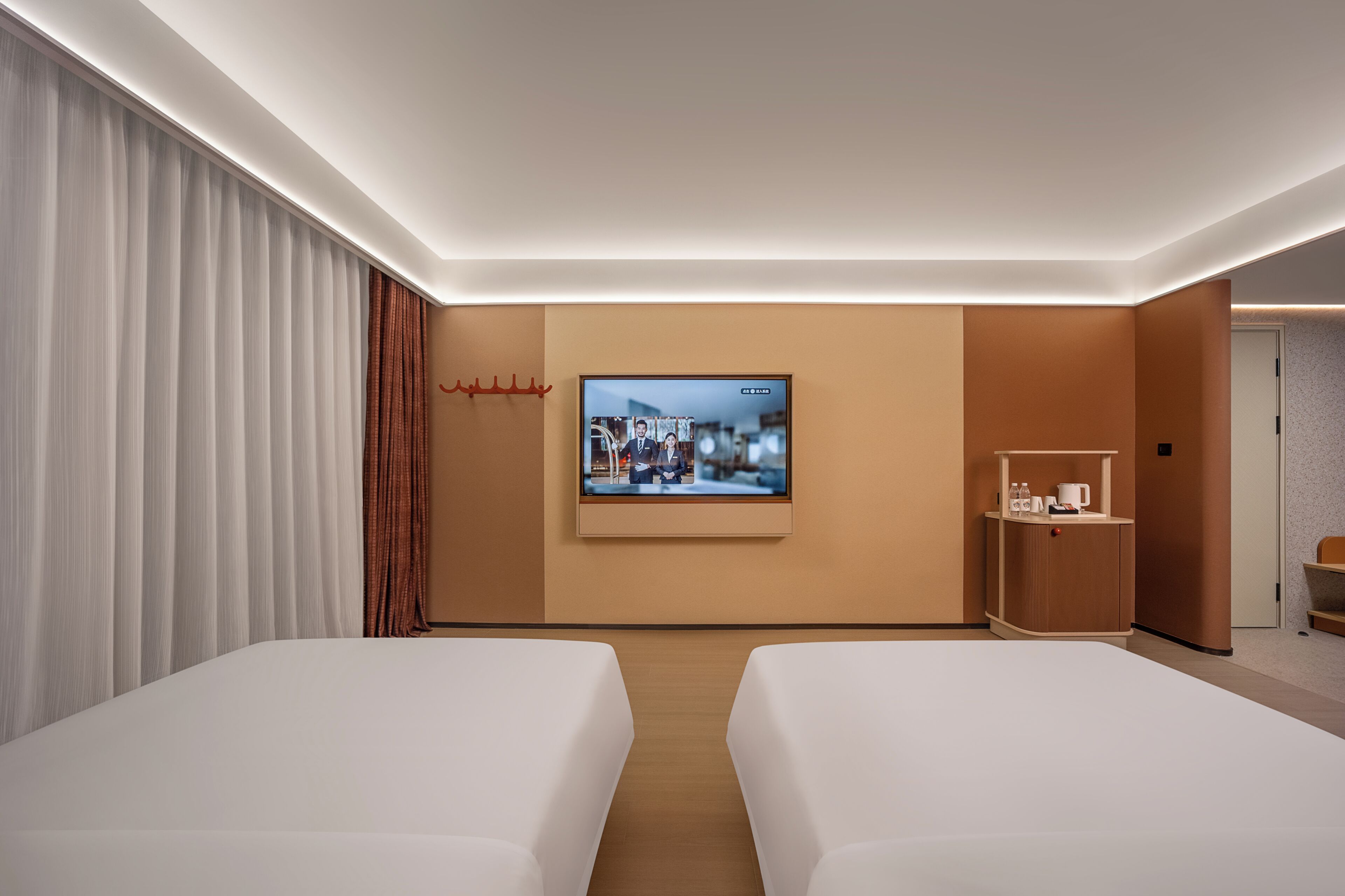 A hotel room with two beds, a television mounted on the wall, a small wooden side table with a cup and a bottle, and a door to the right.