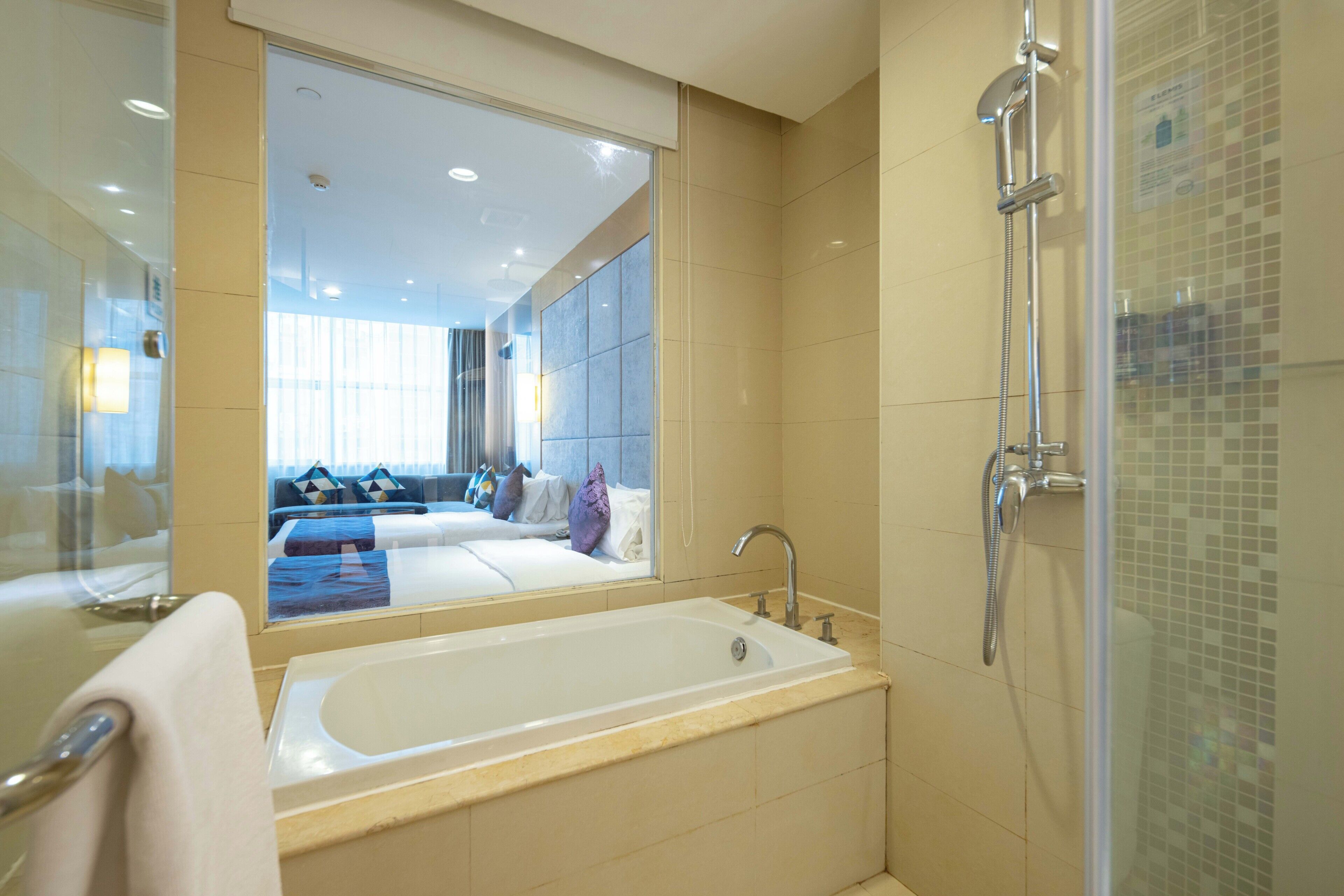 Ein modernes Badezimmer mit einer großen Badewanne, einer Dusche und einem Spiegel, der ein Schlafzimmer mit Bett und Kissen reflektiert.