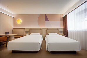 A hotel room with two beds, a desk, a chair, a round wall light, and a large window with curtains.