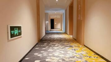 Un pasillo de hotel con piso alfombrado, un detector de incendios a la izquierda y una puerta al final.