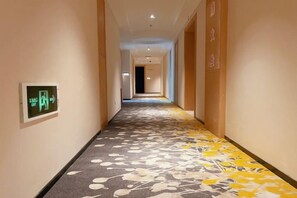 A hotel hallway with a carpeted floor, a fire alarm on the left, and a door at the end.