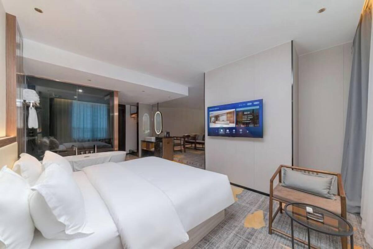 Une chambre d’hôtel moderne dotée d’un grand lit, d’un coin salon avec une petite table, d’un téléviseur fixé au mur et d’une paroi en verre qui sépare la chambre de la salle de bain.