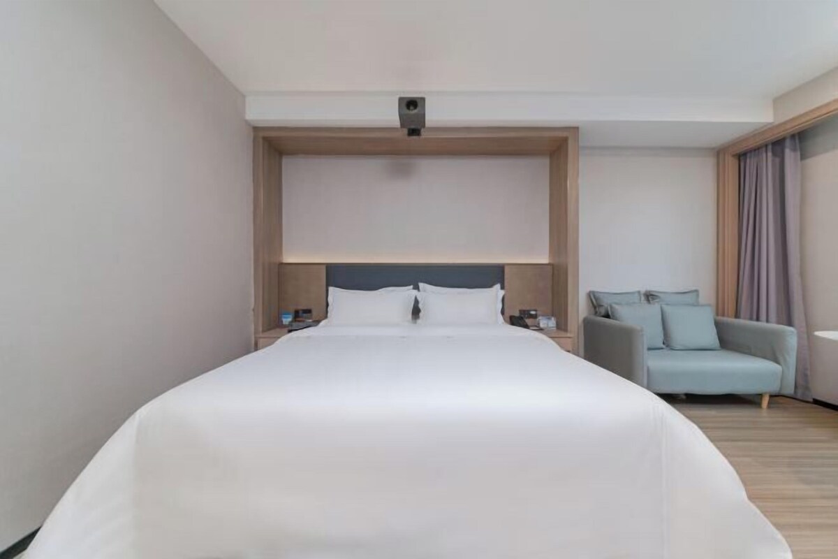 Une chambre d’hôtel moderne avec un grand lit, une tête de lit intégrée, un fauteuil gris et une table de chevet avec une lampe.