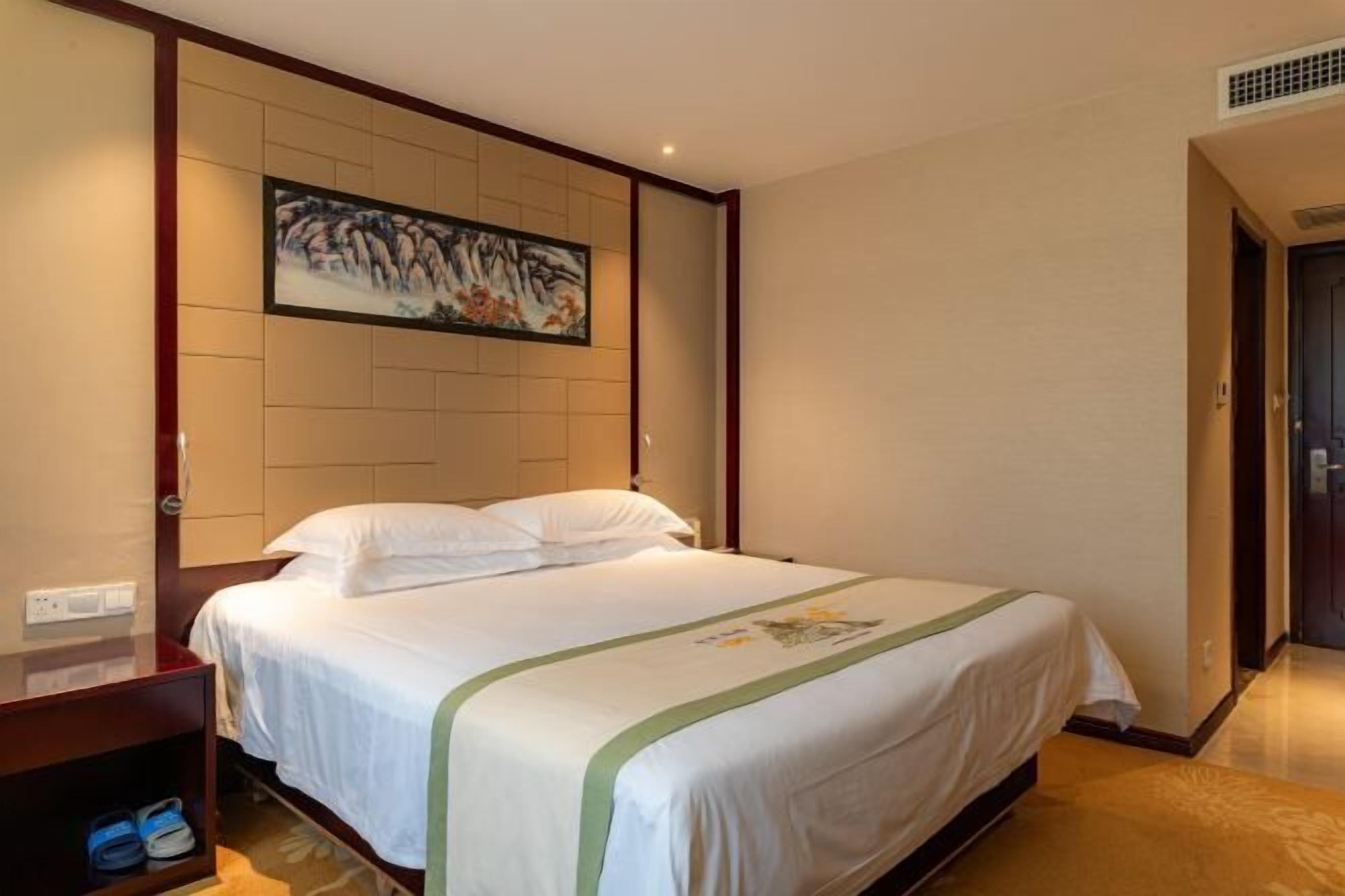 A hotel room with a bed, a nightstand, a pair of slippers, a painting on the wall, and a door to the right.
