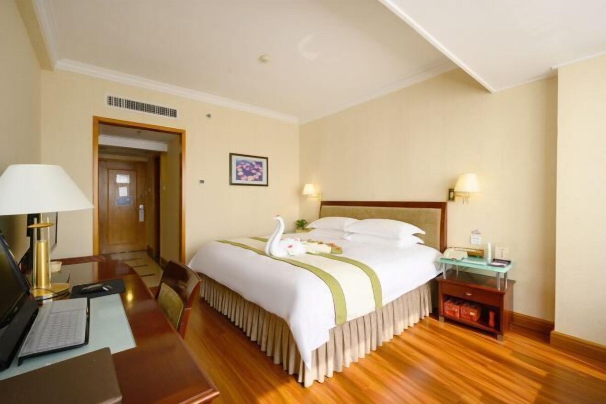A hotel room with a bed, desk, chair, lamp, bedside table, and a door leading to another room.