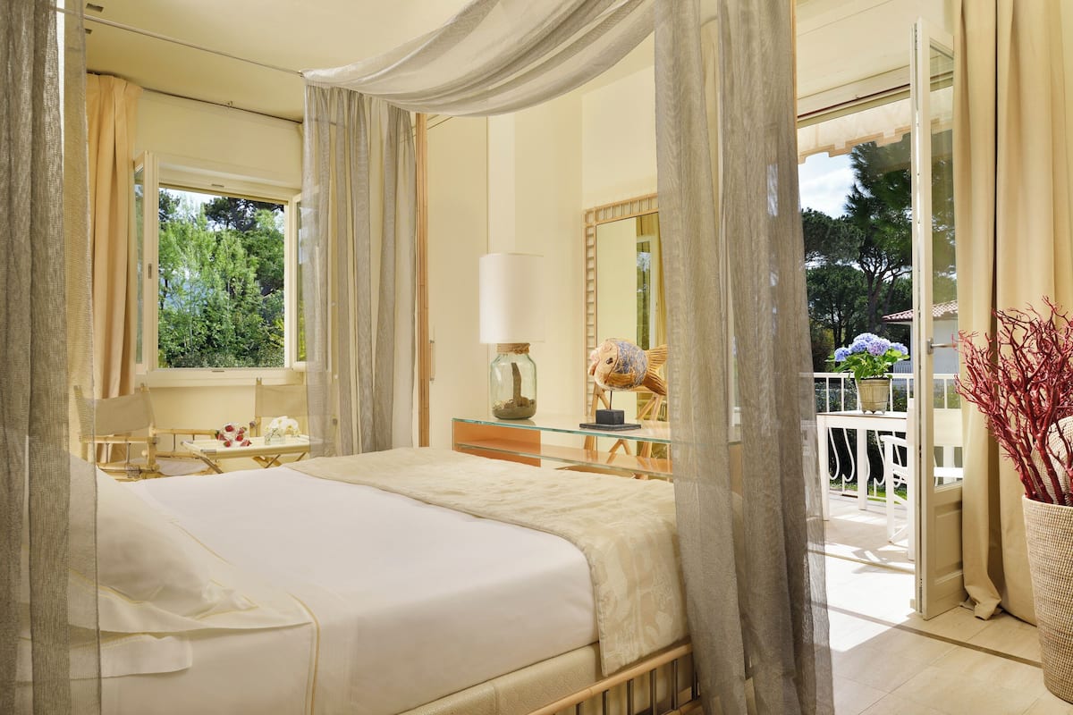 A bedroom with a large bed, a bedside table, a lamp, a mirror, and a view of the outdoor balcony.