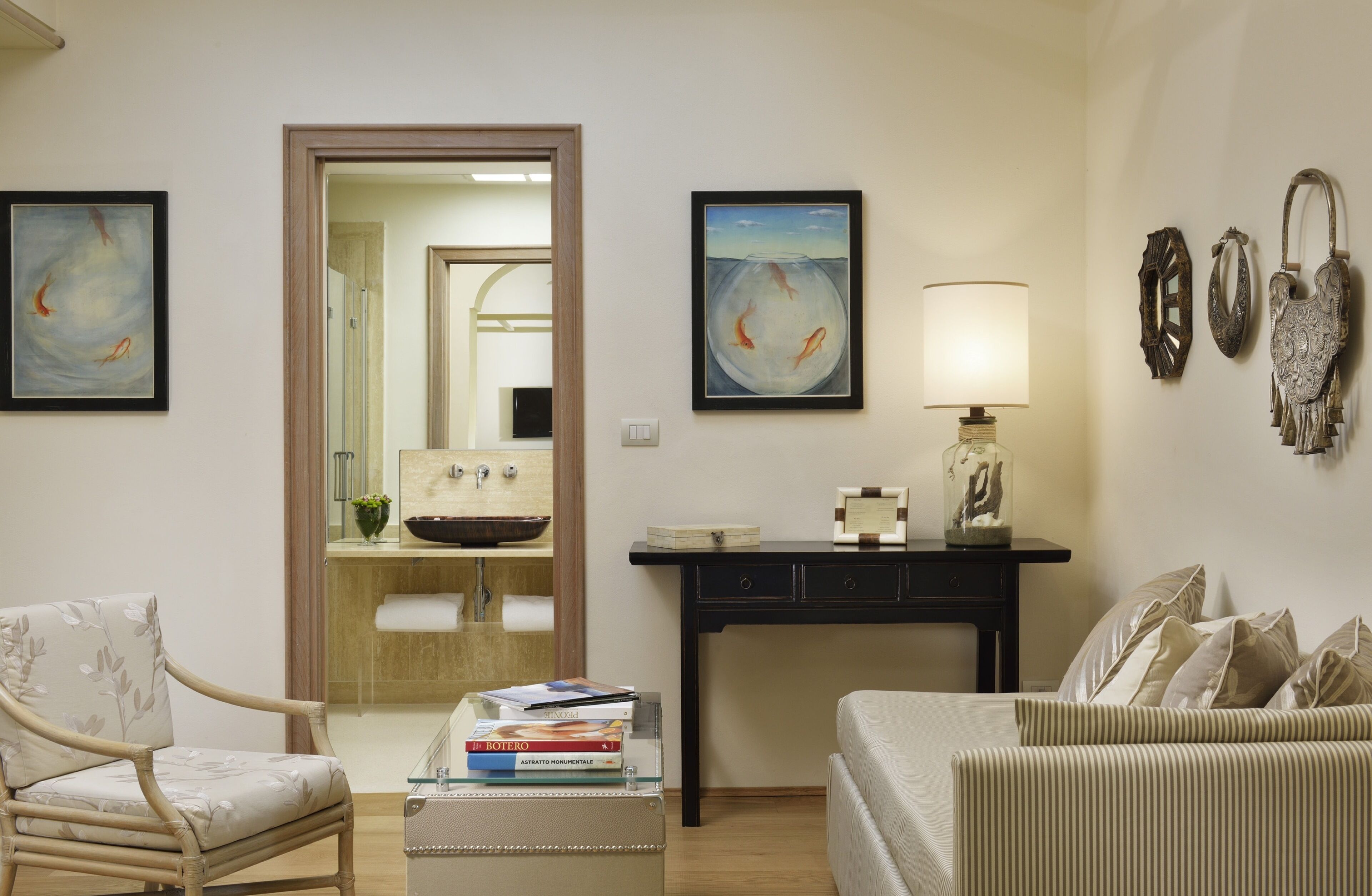 A living room with a sofa, armchair, coffee table, and a desk with a lamp. There are framed artworks on the walls and a mirror in the doorway.