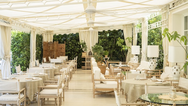 A spacious indoor dining area with round tables and wicker chairs, surrounded by lush greenery and a high ceiling with decorative beams. - Villa Roma Imperiale (Forte dei Marmi)