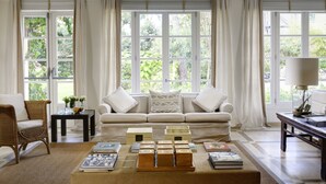 A living room with a white sofa, a coffee table with books, a wicker chair, and large windows with curtains. - Villa Roma Imperiale (Forte dei Marmi)