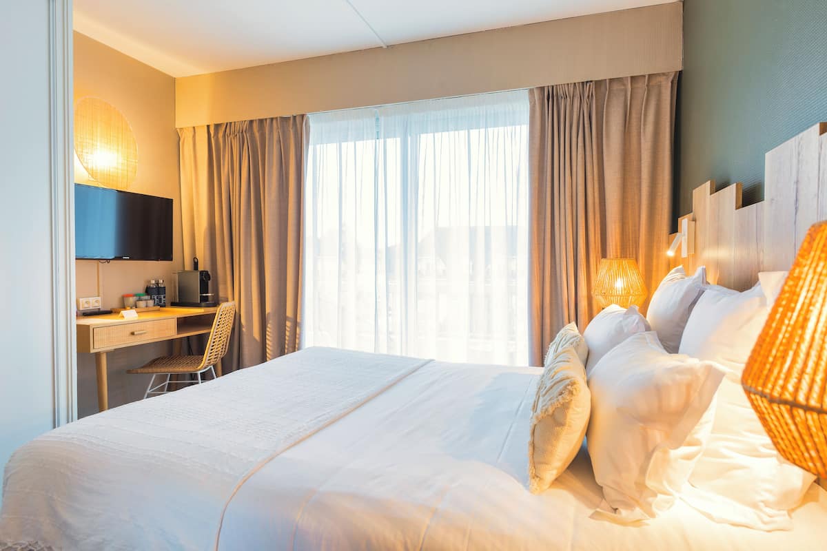 Ein Hotelzimmer mit einem großen Bett, einem Schreibtisch mit Stuhl, einem Fernseher und einem Fenster mit Vorhängen.