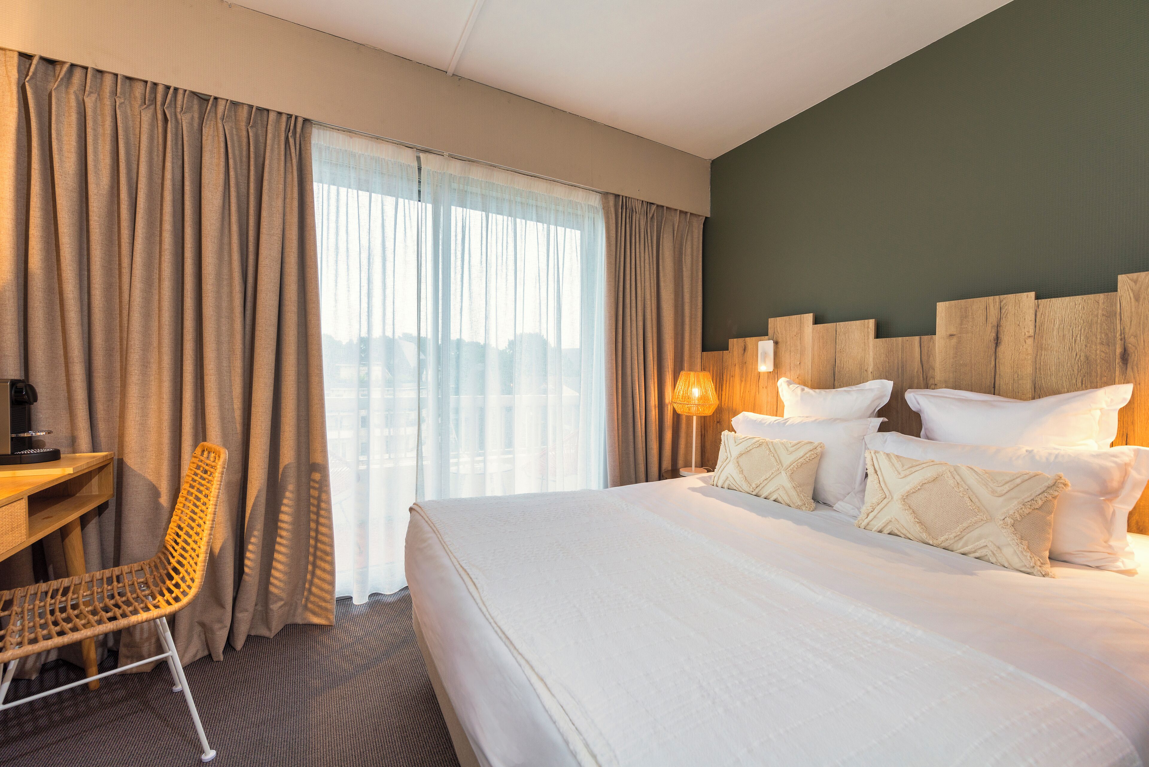 A hotel room with a large bed, a desk, a chair, and a view of the city through sheer curtains.