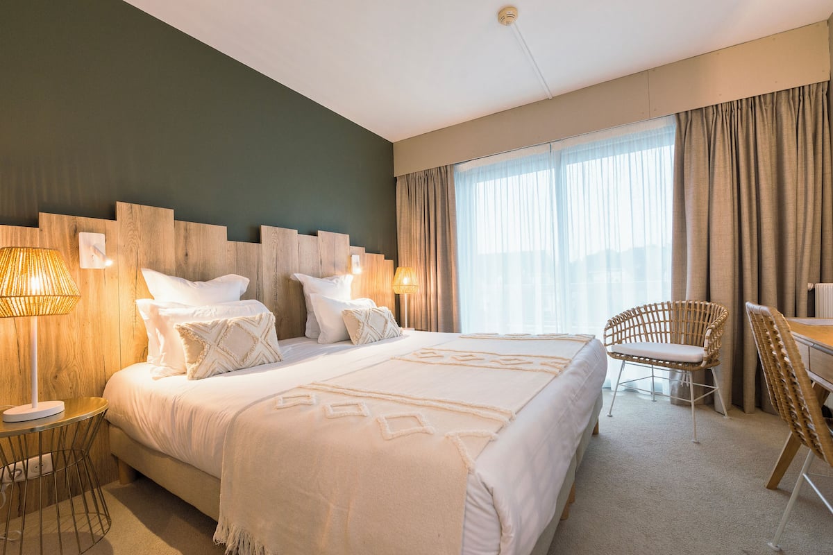 Ein Hotelzimmer mit einem großen Bett, zwei Nachttischlampen, einem Korbsessel, einem Schreibtisch und einem großen Fenster mit Vorhängen.