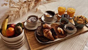 Un plateau de petit-déjeuner disposé sur un lit, avec une variété d’aliments et de boissons, notamment des fruits, des viennoiseries et des boissons.