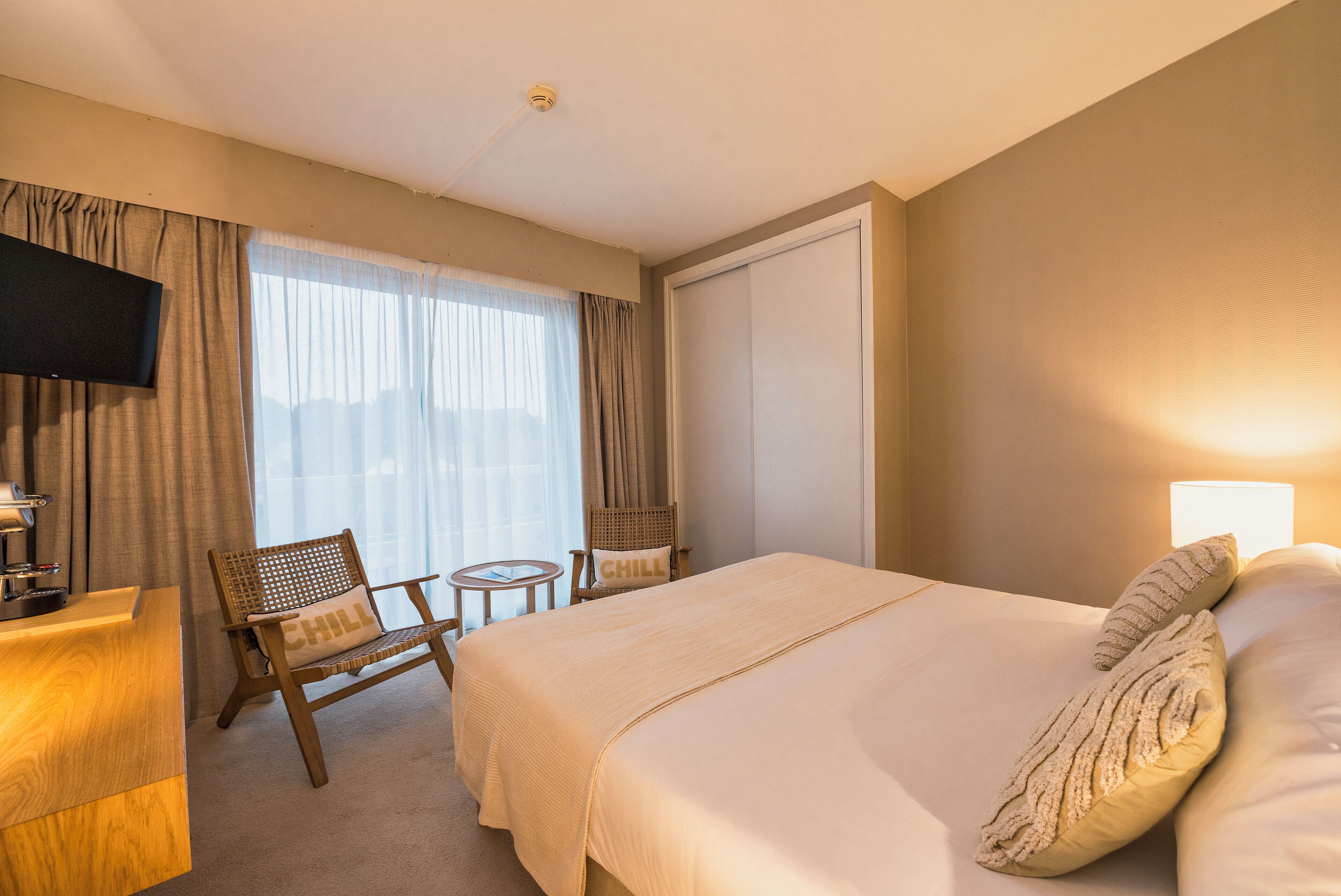 Une chambre d’hôtel avec un lit, un bureau, une chaise, une télévision et une fenêtre avec des rideaux.