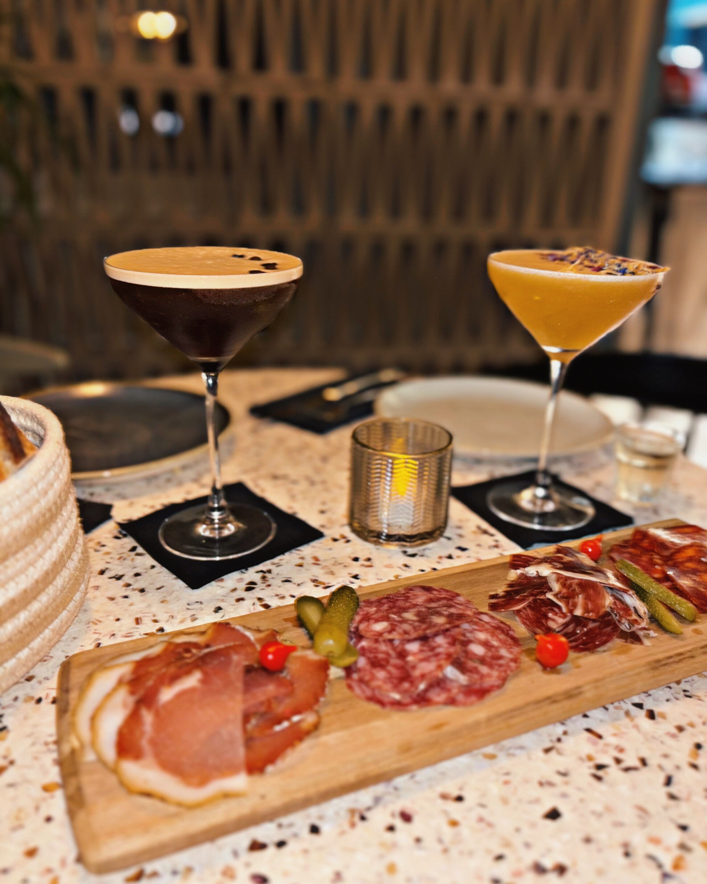 A charcuterie board with various meats and cheeses, two drinks in martini glasses, and a candle on a table.