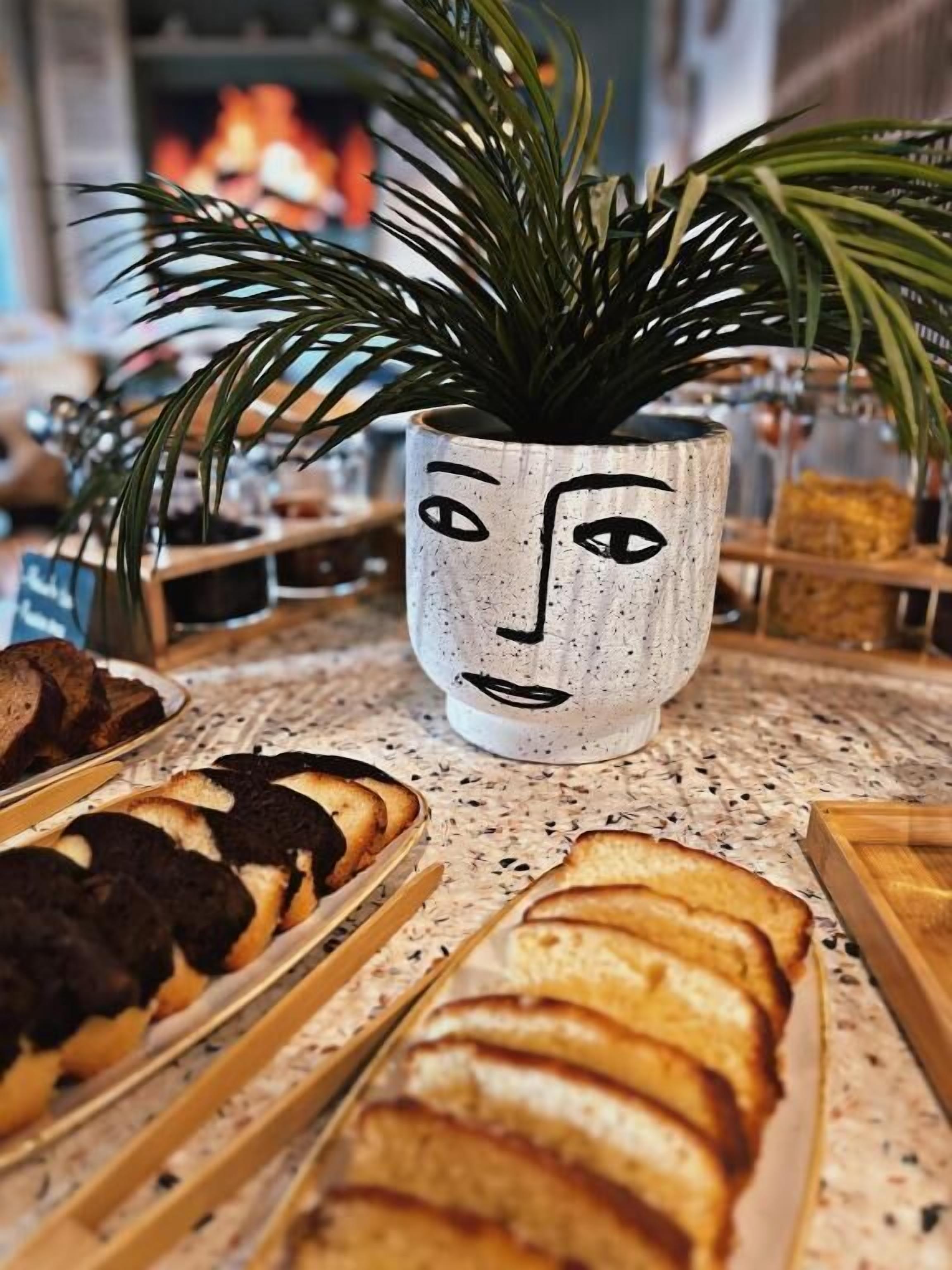 Una mesa con diversos pasteles y pan, una planta en maceta y una taza decorativa con forma de rostro.