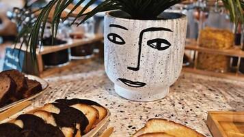 Una mesa con diversos pasteles y pan, una planta en maceta y una taza decorativa con forma de rostro.