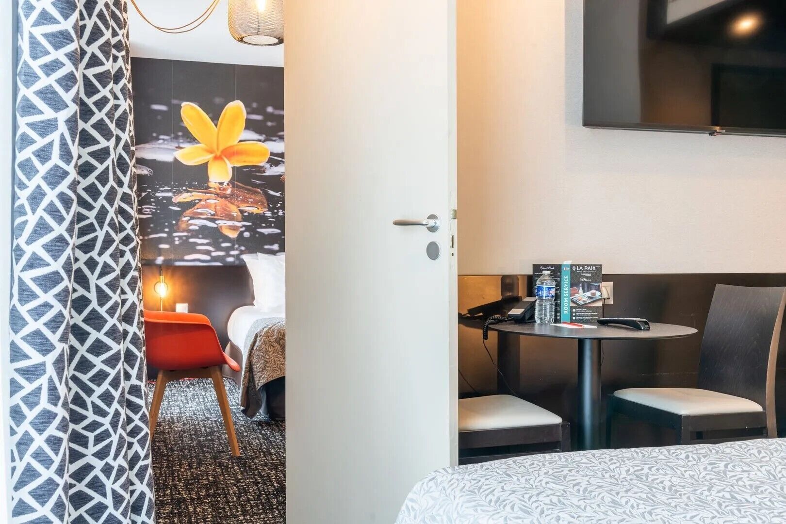 Habitación de hotel con cama, escritorio y silla, una mesita con otra silla, televisor montado en la pared y una puerta con cortina.