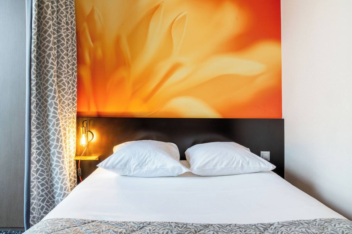 Une chambre d’hôtel avec un lit, des oreillers blancs, une couvre-lit à motifs, un mur orné d’un motif géométrique et un luminaire fixé au mur.