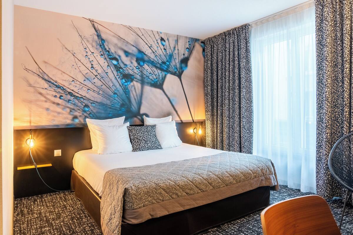 Une chambre d’hôtel avec un grand lit, une lampe fixée au mur et une vue sur l’extérieur à travers des rideaux transparents.