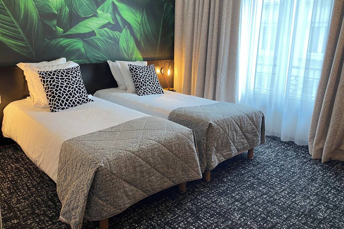 Une chambre d’hôtel avec deux lits, une grande fresque murale représentant des feuilles vertes, une fenêtre avec des rideaux légers et une moquette à motifs.