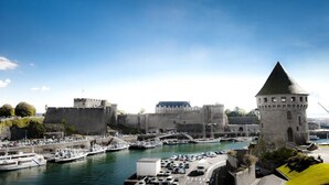Un pueblo costero con un castillo histórico, una torre y un puerto deportivo con barcos y coches.