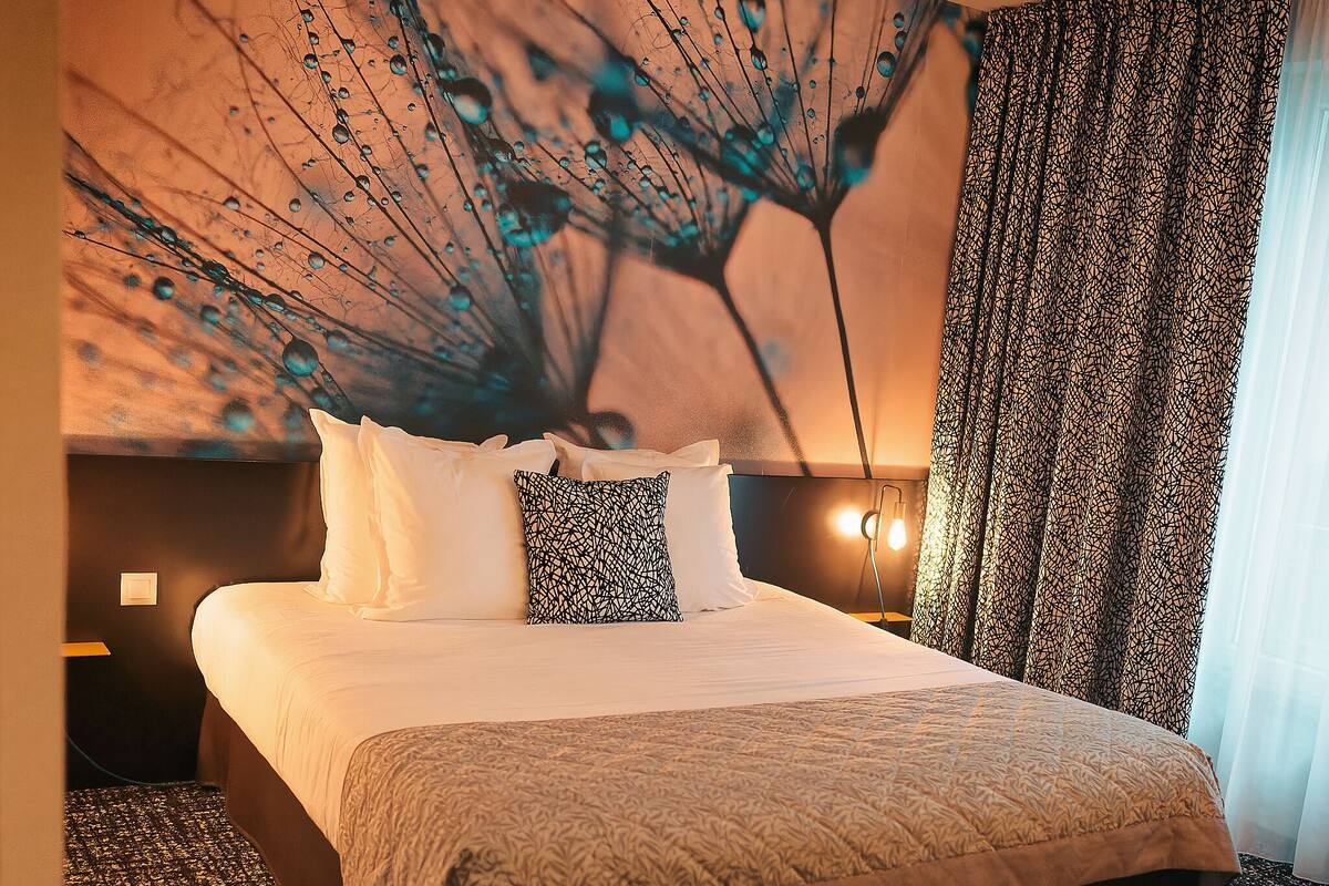 Une chambre d’hôtel avec un grand lit, une tête de lit ornée d’un motif décoratif, une fresque murale et un rideau à motifs.