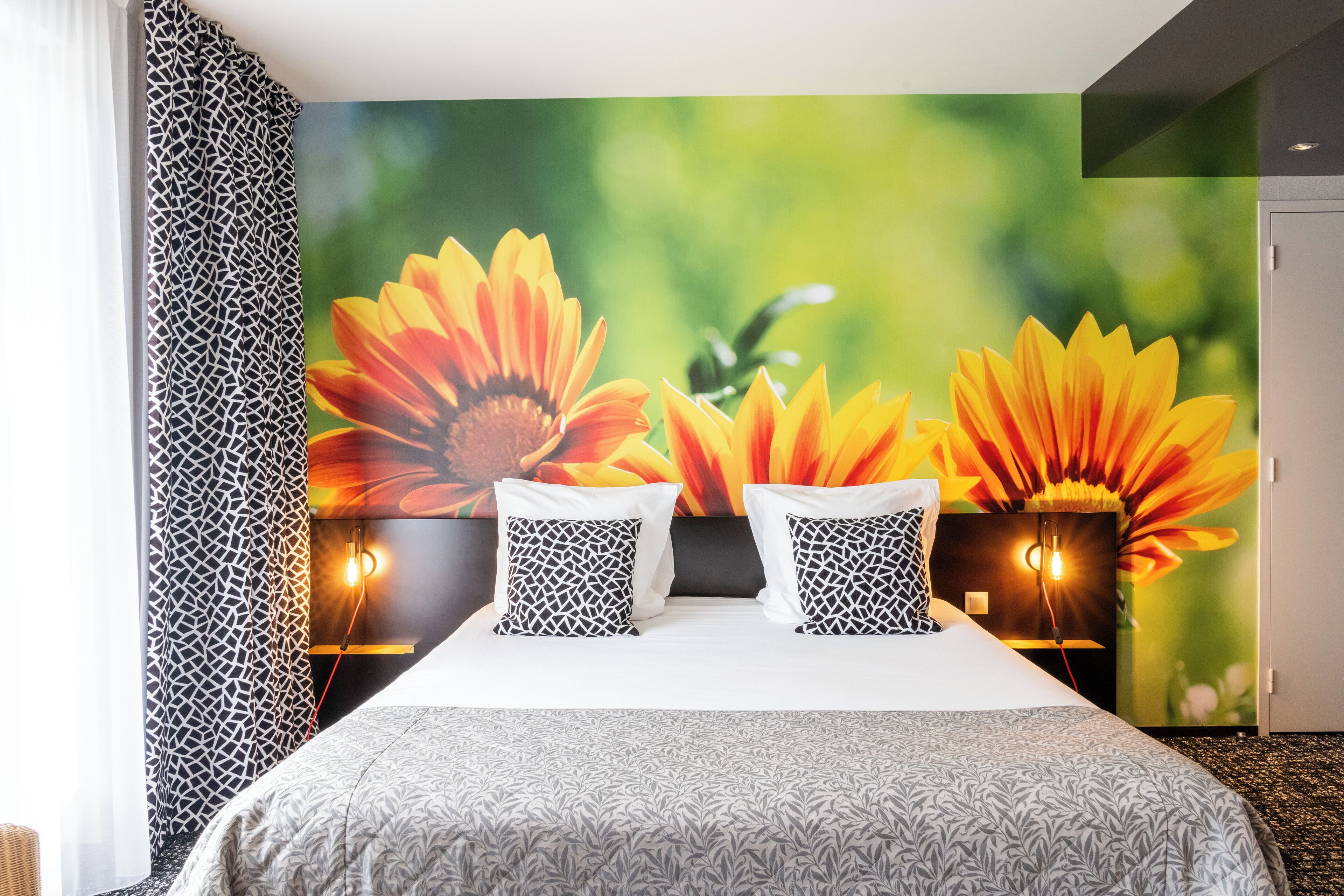 Habitación de hotel con una cama grande, dos almohadas estampadas, un mural floral en la pared y una cortina decorada.