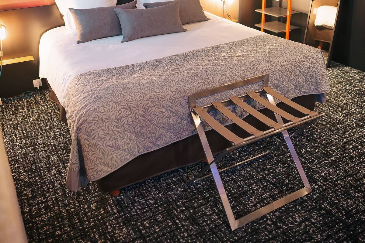Un lit bien fait, avec des oreillers, une table de chevet, une lampe fixée au mur et un tableau.