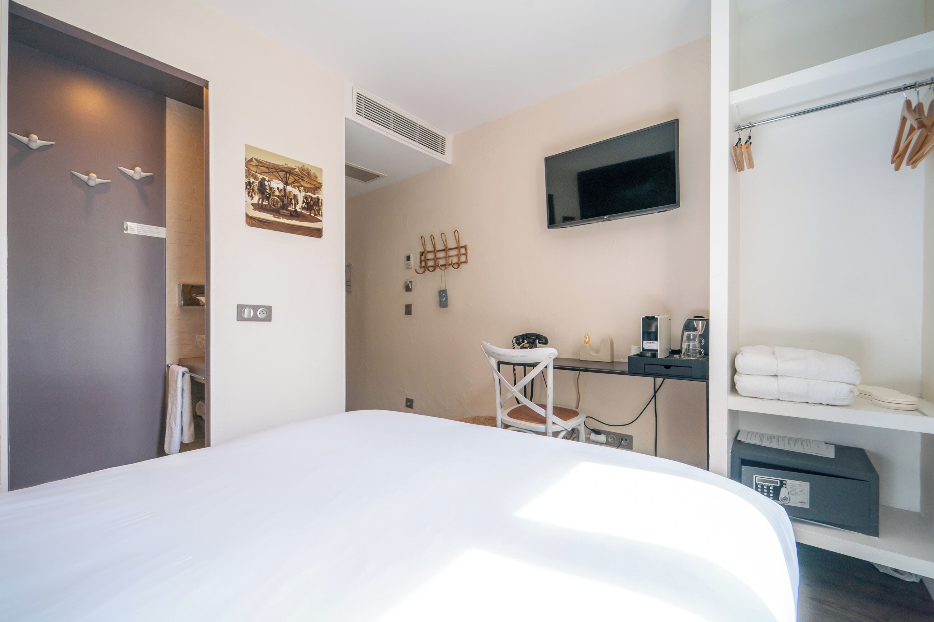 A hotel room with a bed, a desk with a chair, a TV, a coat rack, and a shelf with towels.