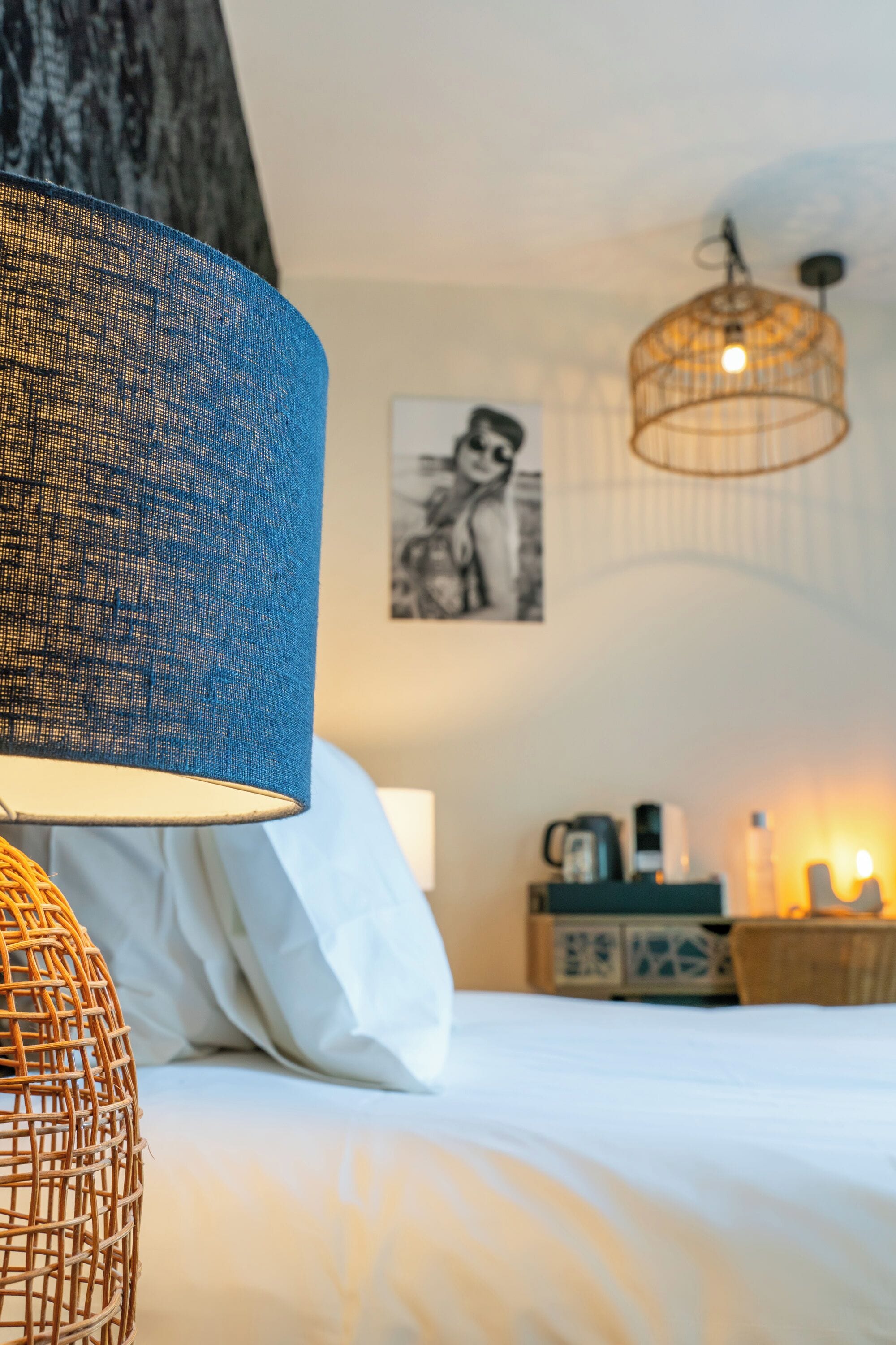 A neatly made bed with a blue and white lamp, a hanging light fixture, and a framed picture on the wall.