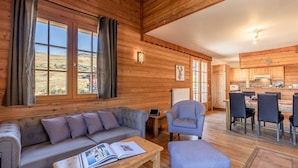 A cozy cabin interior with a wooden interior, a sofa, a dining table with chairs, and a view of the outdoors through large windows. - Madame Vacances Les Chalets de l'Altiport (Huez)