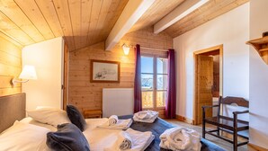 A cozy room with a wooden ceiling, a bed with pillows and towels, a framed picture on the wall, and a window with curtains. - Madame Vacances Les Chalets de l'Altiport (Huez)