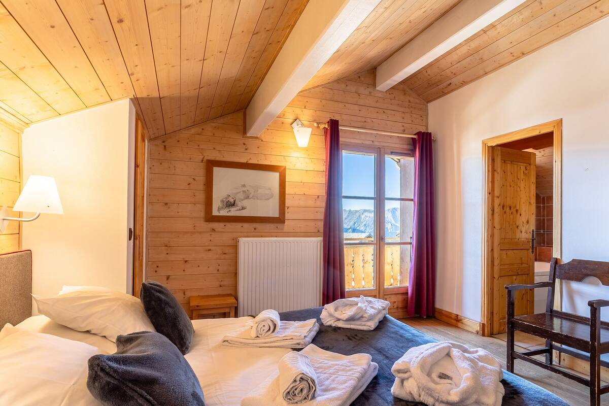Una habitación acogedora con techo de madera, cama con almohadas y toallas, un cuadro enmarcado en la pared y una ventana con cortinas.