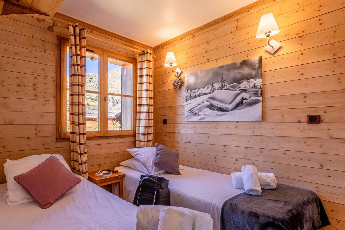 Una habitación acogedora con paredes de madera, dos camas, una ventana con cortinas, un cuadro y una lámpara de pared.