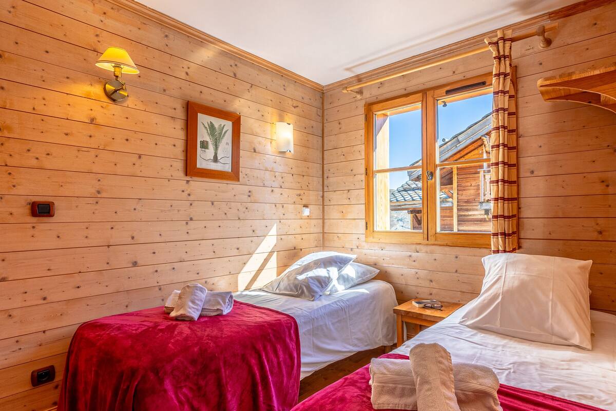 Una habitación de cabaña de madera con dos camas, una ventana con cortinas y una lámpara fijada a la pared.