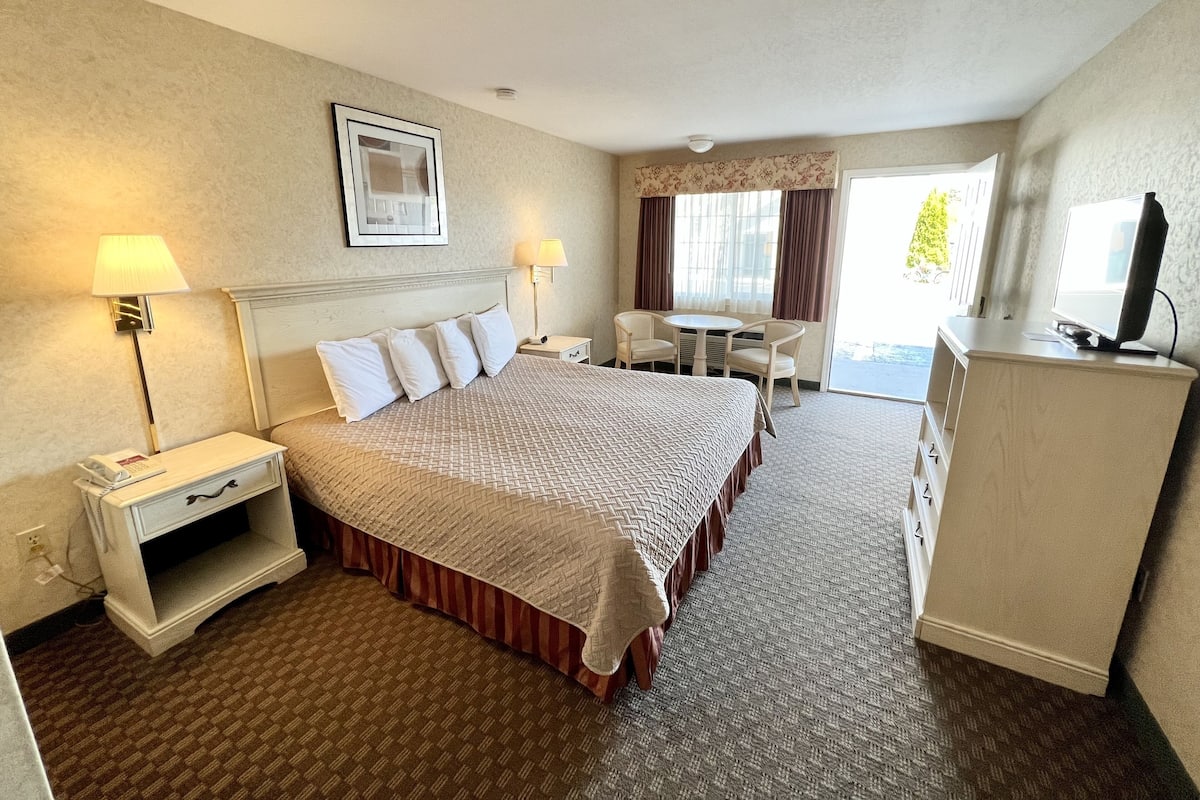 A hotel room with a large bed, bedside tables with lamps, a desk with two chairs, a television on a cabinet, and a window with curtains.