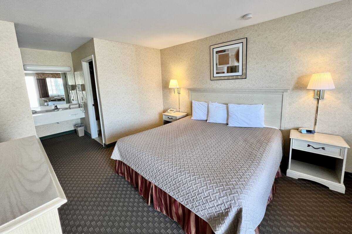 A hotel room with a large bed, a bedside table, a nightstand, a mirror, and a bathroom visible through an open door.