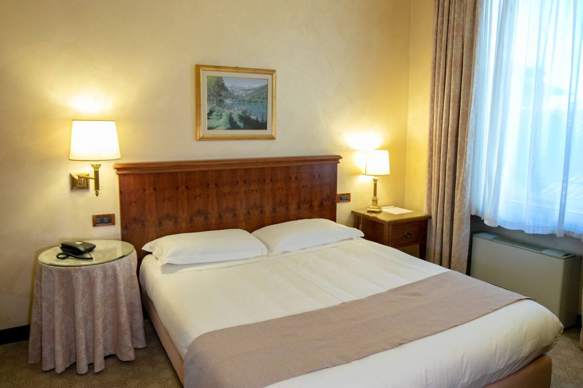 Habitación de hotel con una cama grande, dos mesitas de noche, una lámpara en cada una, un cuadro en la pared y una ventana con cortinas.
