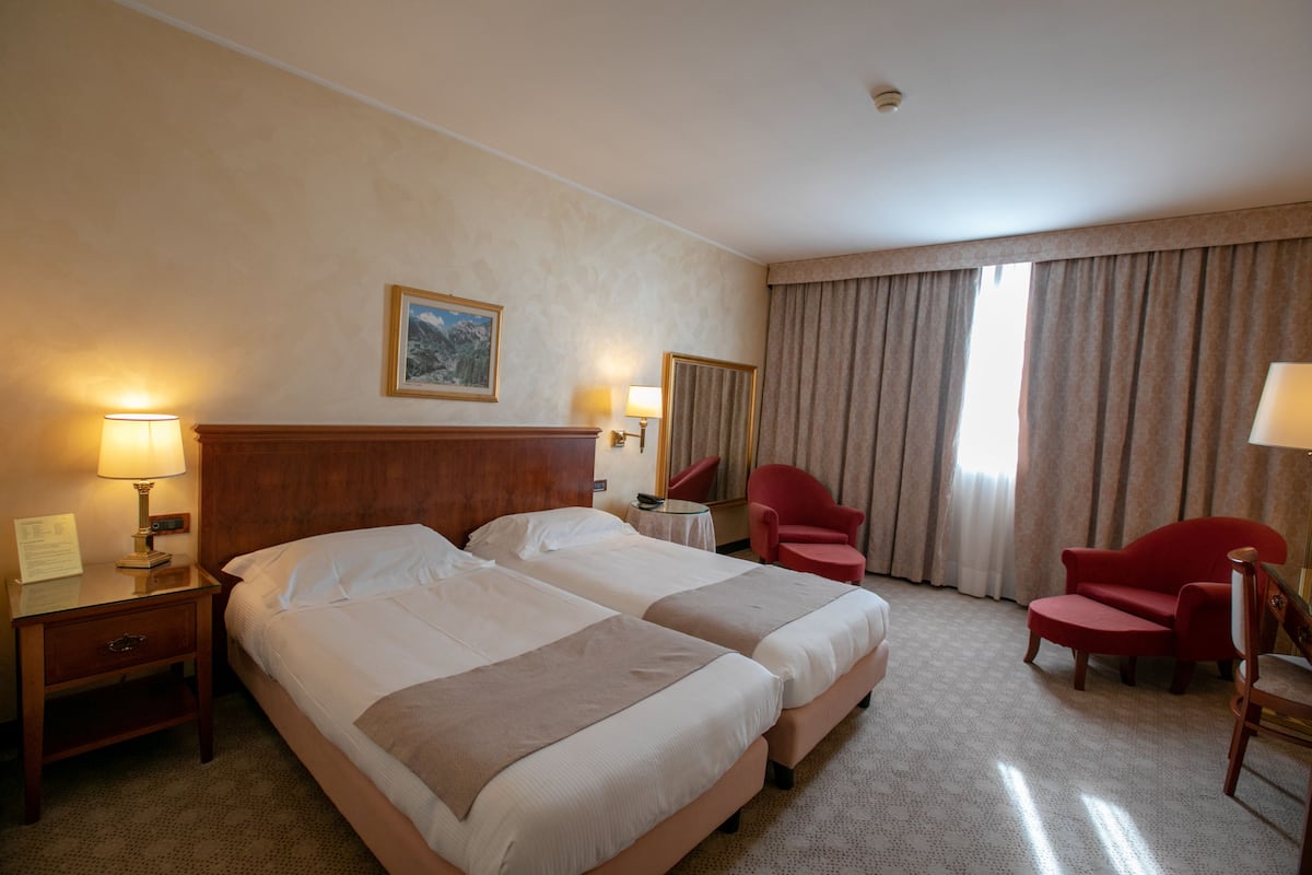 Habitación de hotel con dos camas, cabecera de madera, escritorio con lámpara, una silla y un ventanal con cortinas.
